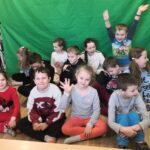 na fotografii widać dzieci siedzące na podłodze na tle zielnego płótna
