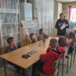 W bibliotecznej sali grupa dzieci siedzi przy stole i słucha z uwagą czytanej im przez bibliotekarza książki o życiu Wandy Rutkiewicz.