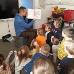 Pan bibliotekarz siedzi na krześle i pokazuje zgromadzonym wokół niego przedszkolakom kolorową ilustrację z książki.