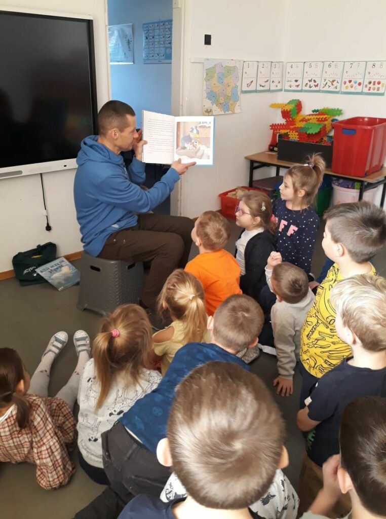 Pan bibliotekarz siedzi na krześle i pokazuje zgromadzonym wokół niego przedszkolakom kolorową ilustrację z książki.