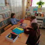 Grupa dzieci siedzi przy bibliotecznym stole i słucha czytanej im przez jedną z dziewczynek książki pt. „O Wilku, który chciał się zakochać”.
