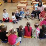 Na podłodze siedzą w kółeczku przedszkolaki. Na małym krzesełku siedzi pani bibliotekarka i czyta dzieciom bajeczkę