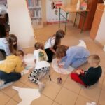 Dzieci siedzą na podłodze i przy pomocy kredek świecowych kolorują wielką papierową głowę kota.
