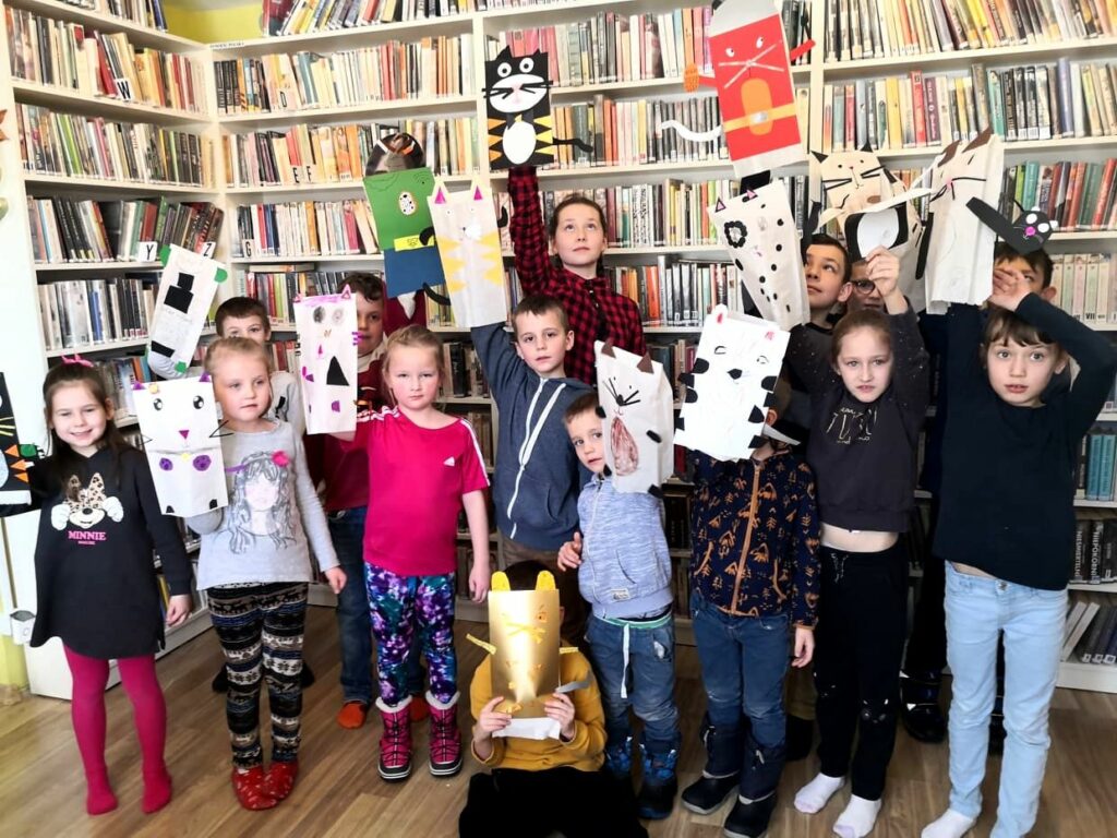 Na zdjęciu widać dzieci stojące na tle regału z książkami. dzieci trzymają w rękach prace plastyczne wykonane przez siebie są to papierowe koty.