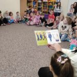 na zdjęciu widać dzieci siedzące na popielatej wykładzinie, siedzą wokół bibliotekarki, która czyta im książkę.