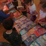 Grupa uczniów stoi przy stolikach na których leży mnóstwo kolorowych książek. Dzieci wybierają i wypożyczają interesujące je lektury.