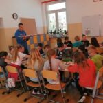 Spora grupa dzieci siedzi przy wielkim stole i koloruje kredkami swoje prace plastyczne. W tym samym czasie stojący obok bibliotekarz czyta im zabawne opowiadanie Joanny Krzyżanek.