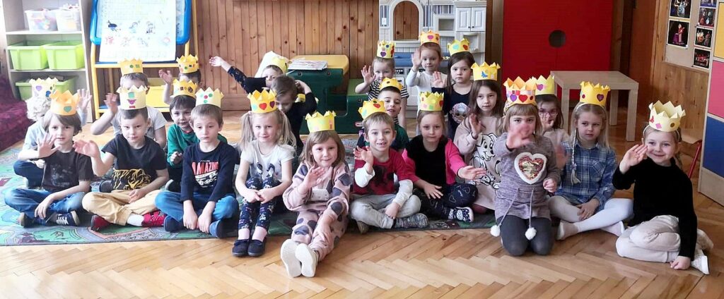 na zdjęciu widać dzieci siedzące na podłodze w sali przedszkolnej. Dzieci na głowie mają papierowe korony.