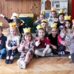 na zdjęciu widać dzieci siedzące na podłodze w sali przedszkolnej. Dzieci na głowie mają papierowe korony.