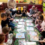 na zdjęciu widać dzieci siedzące w kole, w środku widoczne są widać ilustracje z bajek, wśród dzieci siedzi bibliotekarka trzymające białe karteczki.