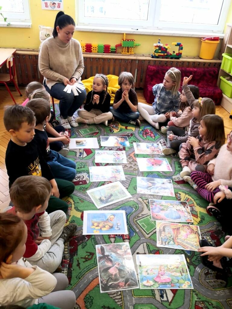 na zdjęciu widać dzieci siedzące w kole, w środku widoczne są widać ilustracje z bajek, wśród dzieci siedzi bibliotekarka trzymające białe karteczki.