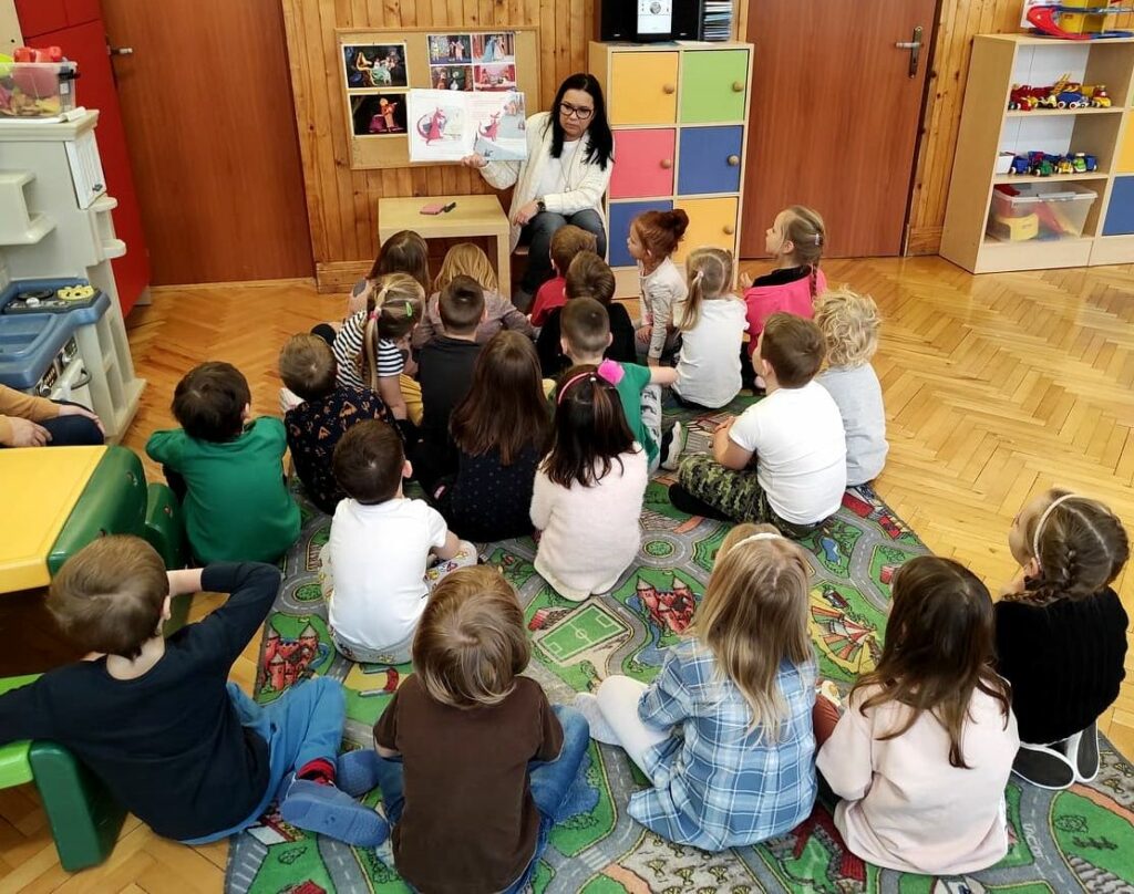na zdjęciu widać siedzące na podłodze patrzą w stronę bibliotekarki, która trzyma bajkę.