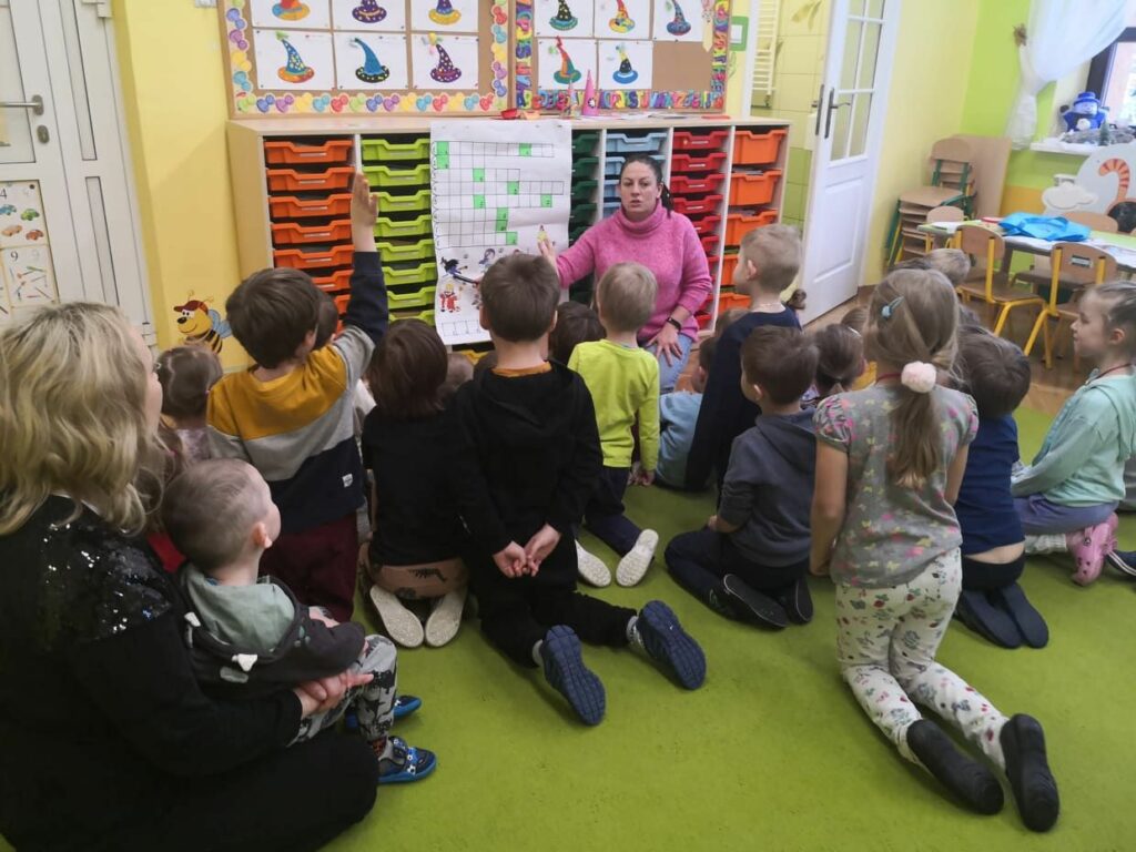 na zdjęciu widać bibliotekarkę, która siedzi przed dziećmi w wieku przedszkolnym wspólnie pozawiązują kolorową krzyżówkę.