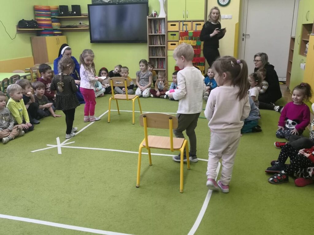 na zdjęciu widać zieloną wykładzinę siedzące na niej przedszkolaki kilkoro dzieci chodzi z talerzykami wzdłuż zaznaczonej linii.