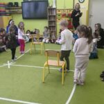 na zdjęciu widać zieloną wykładzinę siedzące na niej przedszkolaki kilkoro dzieci chodzi z talerzykami wzdłuż zaznaczonej linii.
