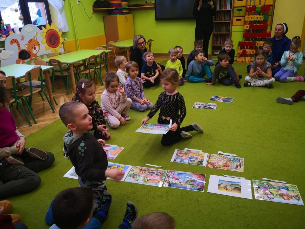 na zdjęciu widać kolorową sale przedszkolną, zieloną wykładzinę na której siedzą dzieci i układają kolorowe obrazki - ilustracje z bajek.