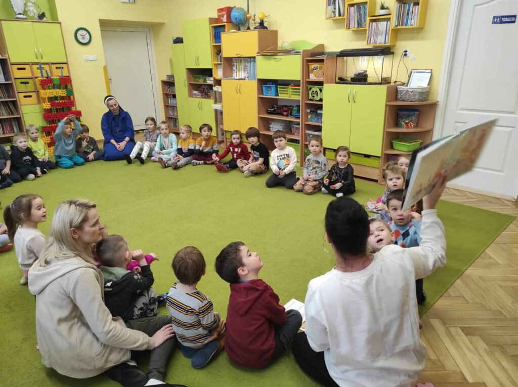 na zdjęciu widać kolorową salę przedszkolną, zieloną wykładzinę i dzieci siedzące w kole słuchające bibliotekarki, która pokazuje im książkę.