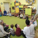 na zdjęciu widać kolorową salę przedszkolną, zieloną wykładzinę i dzieci siedzące w kole słuchające bibliotekarki, która pokazuje im książkę.