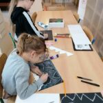 Dzieci pracują przy stole rysując na kartkach pastelami i wycinając z papieru szablony niedźwiadków polarnych.
