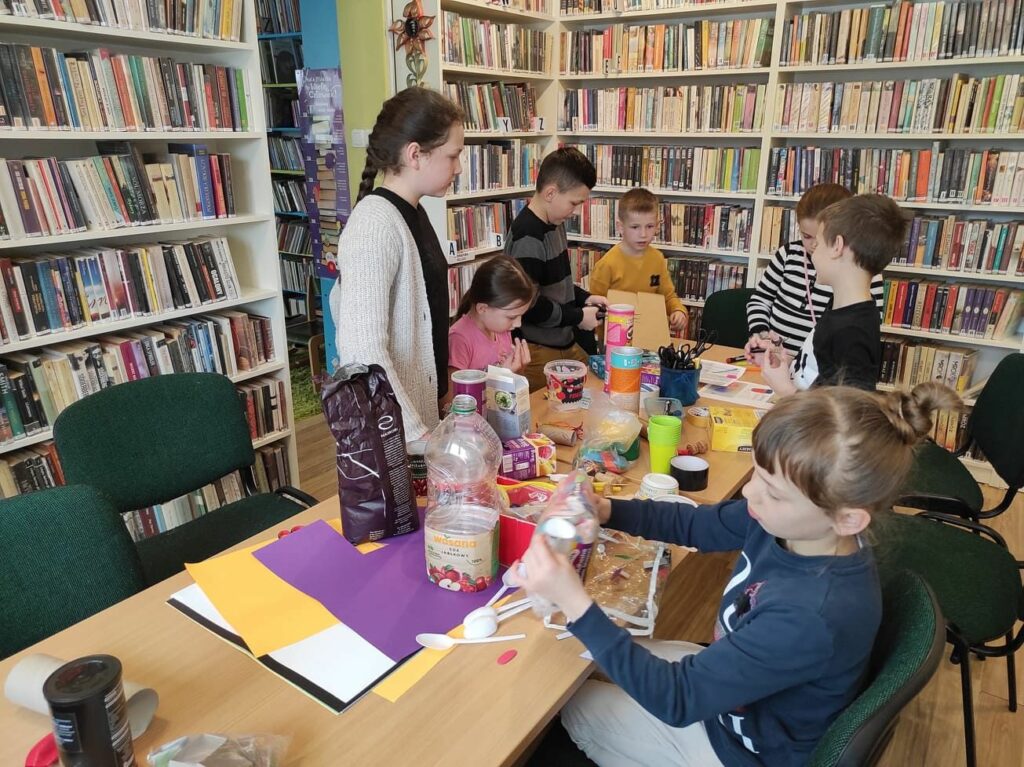 na zdjęciu widać salę biblioteczną, regały z książkami, na środku sali stoi stół, wokół stołu siedzą dzieci, wykonują pracę plastyczną