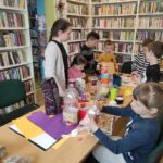 na zdjęciu widać salę biblioteczną, regały z książkami, na środku sali stoi stół, wokół stołu siedzą dzieci, wykonują pracę plastyczną