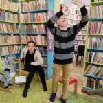 na zdjęciu widać salę biblioteczną, regały z książkami, widać dwoje dzieci, dziewczynka siedzi na krześle, chłopiec stoi na środku sali