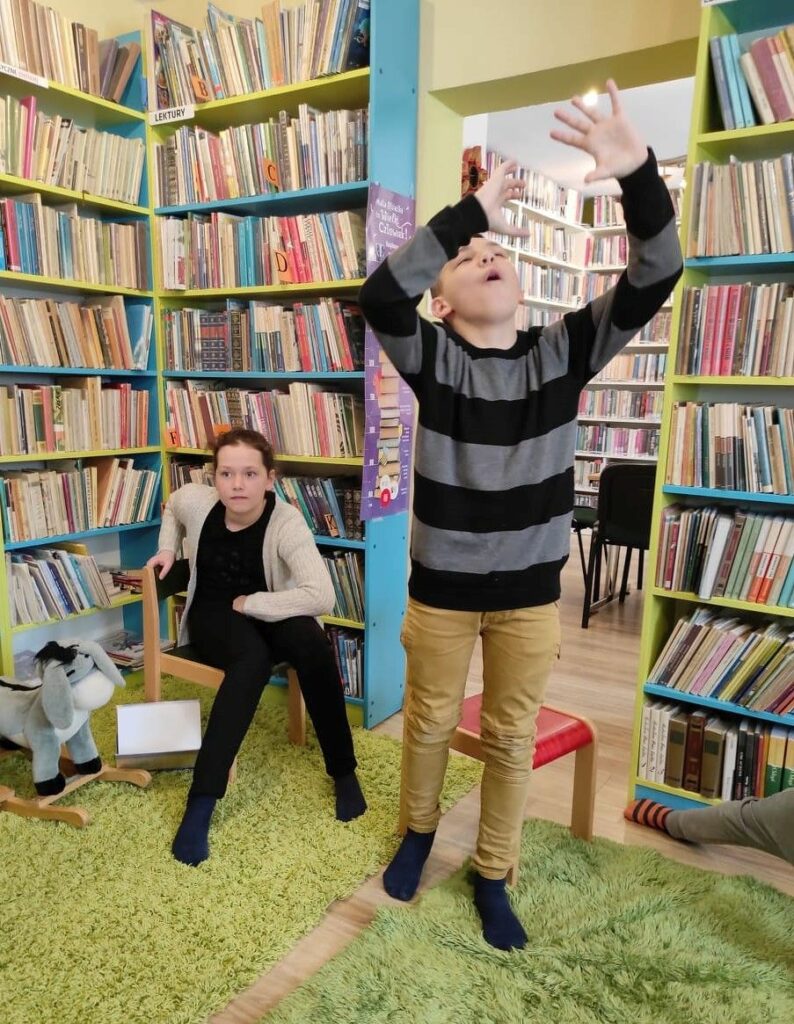 na zdjęciu widać salę biblioteczną, regały z książkami, widać dwoje dzieci, dziewczynka siedzi na krześle, chłopiec stoi na środku sali