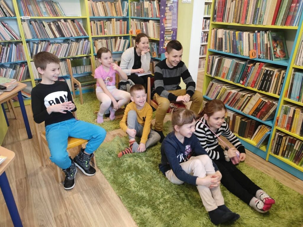 na zdjęciu widać salę biblioteczną, regały z książkami, na środku sali na dywanie siedzą dzieci