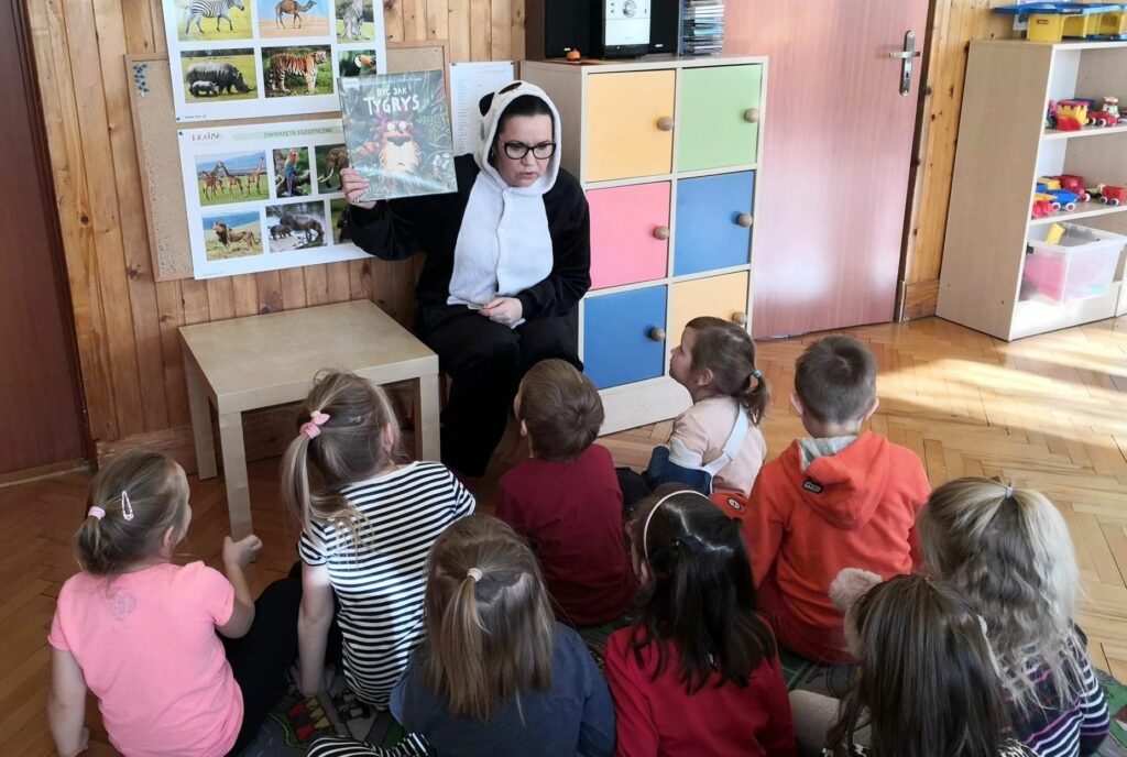 Na zdjęciu widać siedząca na krześle bibliotekarkę w stroju pandy pokazującą siedzącym przed nią dzieciom książkę.