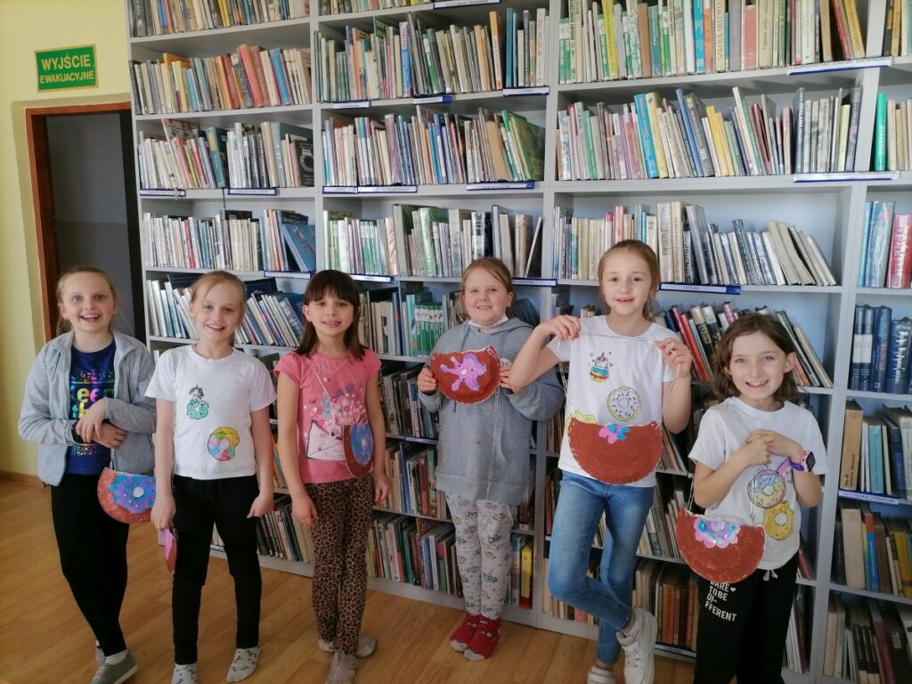 Na zdjęciu widoczne są dzieci uczestniczące w zajęciach zorganizowanych przez Bibliotekę w Tyliczu przy współpracy Centrum Kultury oraz Świetlicy "Przystań" z okazji Tłustego Czwartku dzieci prezentują wykonane przez siebie koszulki z rysunkami pączków i donut'ów oraz torebki w kształcie pączków wykonane z papierowych talerzyków. W tle regały z książkami.