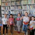 Na zdjęciu widoczne są dzieci uczestniczące w zajęciach zorganizowanych przez Bibliotekę w Tyliczu przy współpracy Centrum Kultury oraz Świetlicy "Przystań" z okazji Tłustego Czwartku dzieci prezentują wykonane przez siebie koszulki z rysunkami pączków i donut'ów oraz torebki w kształcie pączków wykonane z papierowych talerzyków. W tle regały z książkami.