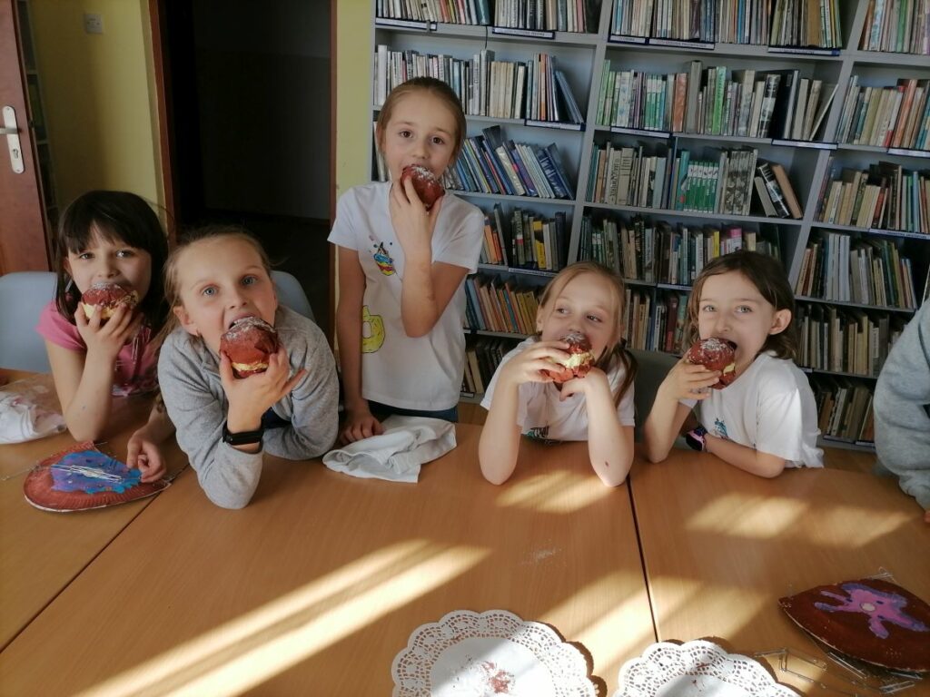 Na zdjęciu widoczne są dzieci uczestniczące w zajęciach zorganizowanych przez Bibliotekę w Tyliczu przy współpracy Centrum Kultury oraz Świetlicy "Przystań" z okazji Tłustego Czwartku dzieci udają że zjadają wykonane przez siebie pączki z papieru. W tle regały z książkami.