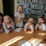 Na zdjęciu widoczne są dzieci uczestniczące w zajęciach zorganizowanych przez Bibliotekę w Tyliczu przy współpracy Centrum Kultury oraz Świetlicy "Przystań" z okazji Tłustego Czwartku dzieci udają że zjadają wykonane przez siebie pączki z papieru. W tle regały z książkami.