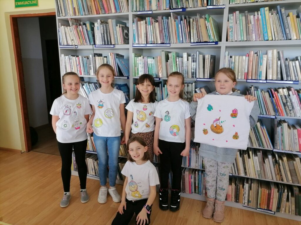 Na zdjęciu widoczne są dzieci uczestniczące w zajęciach zorganizowanych przez Bibliotekę w Tyliczu przy współpracy Centrum Kultury oraz Świetlicy "Przystań" z okazji Tłustego Czwartku dzieci prezentują wykonane przez siebie koszulki z rysunkami pączków i donut'ów. W tle regały z książkami.
