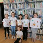 Na zdjęciu widoczne są dzieci uczestniczące w zajęciach zorganizowanych przez Bibliotekę w Tyliczu przy współpracy Centrum Kultury oraz Świetlicy "Przystań" z okazji Tłustego Czwartku dzieci prezentują wykonane przez siebie koszulki z rysunkami pączków i donut'ów. W tle regały z książkami.