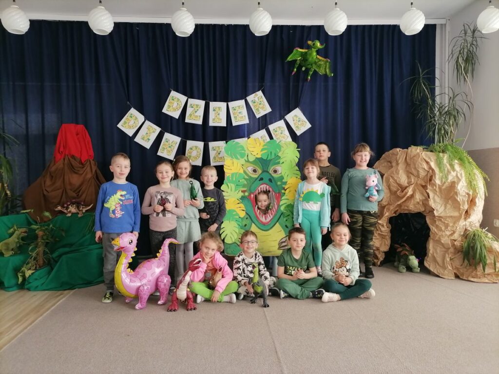 Na zdjęciu widoczna jest grupa przedszkolaków pozująca do wspólnego zdjęcia podczas zajęć z okazji Dnia Dinozaura. Siedmioro dzieci stoi, czworo siedzi na dywanie, jedna dziewczynka pozuje w stworzonej foto-budce - "paszczy dinozaura". Za dziećmi widoczne jest granatowe tło na którym zawieszony jest napis "Dzień Dinozaura" oraz dmuchany pterodaktyl. Po lewej stronie jest ustawiona atrapa wulkanu i pluszowe dinozaury. Z prawej strony jest ustawiona atrapa groty i pluszowy dinozaur. Większość dzieci trzyma w dłoniach plastikowe lub pluszowe dinozaury