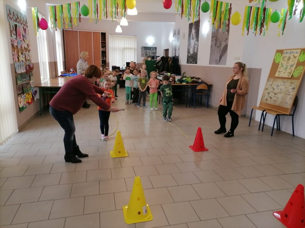 Na zdjęciu widoczna jest grupa przedszkolaków podzielona na dwie drużyny oraz trzy opiekunki. Przedszkolaki są ustawione w dwóch rzędach i biorą udział w zabawie "dinozaurowy tor przeszkód", gdzie w pierwszej kolejności muszą sprawnie przejść po rozciągniętej na podłodze linie, a następnie z trzymanym na łyżce plastikowym jajkiem przemierzyć trasę wokół ustawionych pachołków.
