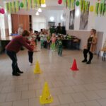Na zdjęciu widoczna jest grupa przedszkolaków podzielona na dwie drużyny oraz trzy opiekunki. Przedszkolaki są ustawione w dwóch rzędach i biorą udział w zabawie "dinozaurowy tor przeszkód", gdzie w pierwszej kolejności muszą sprawnie przejść po rozciągniętej na podłodze linie, a następnie z trzymanym na łyżce plastikowym jajkiem przemierzyć trasę wokół ustawionych pachołków.
