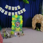 na zdjęciu widoczny jest chłopiec pozujący w specjalnie przygotowanej foto-budce "Paszczy dinozaura" Za foto-budką widoczne jest granatowe tło i przywieszony do niego napi dzień Dinozaura. Przed foto-budką ustawione są figurki dinozaurów. Z prawej strony widoczna jest atrapa jaskini z pluszowym dinozaurem.