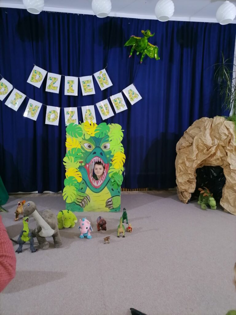 na zdjęciu widoczny jest chłopiec pozujący w specjalnie przygotowanej foto-budce "Paszczy dinozaura" Za foto-budką widoczne jest granatowe tło i przywieszony do niego napi dzień Dinozaura. Przed foto-budką ustawione są figurki dinozaurów. Z prawej strony widoczna jest atrapa jaskini z pluszowym dinozaurem.