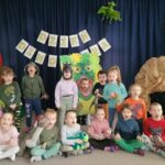 Na zdjęciu widoczna jest grupa przedszkolaków pozująca do wspólnego zdjęcia podczas zajęć z okazji Dnia Dinozaura. Pięcioro dzieci stoi, siedmioro siedzi na dywanie, jeden chłopiec pozuje w stworzonej foto-budce - "paszczy dinozaura". Za dziećmi widoczne jest granatowe tło na którym zawieszony jest napis "Dzień Dinozaura" oraz dmuchany pterodaktyl. Po lewej stronie jest ustawiona atrapa wulkanu i pluszowe dinozaury. Z prawej strony jest ustawiona atrapa groty i pluszowy dinozaur. Większość dzieci trzyma w dłoniach plastikowe lub pluszowe dinozaury