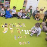 na zdjęciu widać dzieci siedzące na zielonej wykładzinie i przyglądające się fotografią przedstawiającym dzikie zwierzęta.