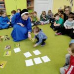 na zdjęciu widać dzieci wraz z opiekunkami, w tym siostrę zakonną siedzących na zielonej wykładzinie i układające ilustracje przedstawiające dzikie zwierzęta.