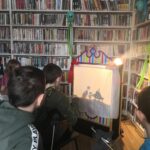 na zdjęciu widać salę biblioteczną, regały z książkami, przy regałach z książkami stoi konstrukcja tworzącą scenę teatralną, obok świeci lampa, dzieci tworzą teatr cieni