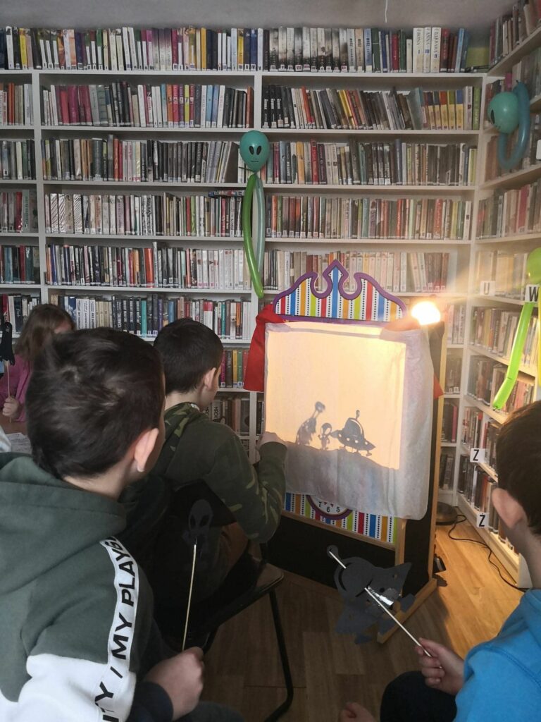 na zdjęciu widać salę biblioteczną, regały z książkami, przy regałach z książkami stoi konstrukcja tworzącą scenę teatralną, obok świeci lampa, dzieci tworzą teatr cieni