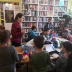 na zdjęciu widać salę biblioteczną, regały z książkami, na środku sali stoi stół , na stole leżą materiały plastyczne, dzieci siedzą wokół stołu wykonują pracę plastyczną