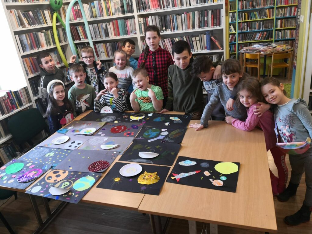 na zdjęciu widać salę biblioteczną, regały z książkami, na środku sali stoi stół , na stole leża wykonane prace plastyczne dzieci, wokół stołu stoją dzieci
