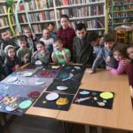 na zdjęciu widać salę biblioteczną, regały z książkami, na środku sali stoi stół , na stole leża wykonane prace plastyczne dzieci, wokół stołu stoją dzieci