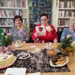 na zdjęciu widać Panie seniorki siedzące za stołem. Jedna z Pań czyta życzenia z kartki, którą trzyma w rękach.