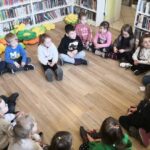 na zdjęciu widać dzieci siedzące na podłodze w sali bibliotecznej wraz z bibliotekarką, która przekazuje im niebieski balonik napełniony wodą.